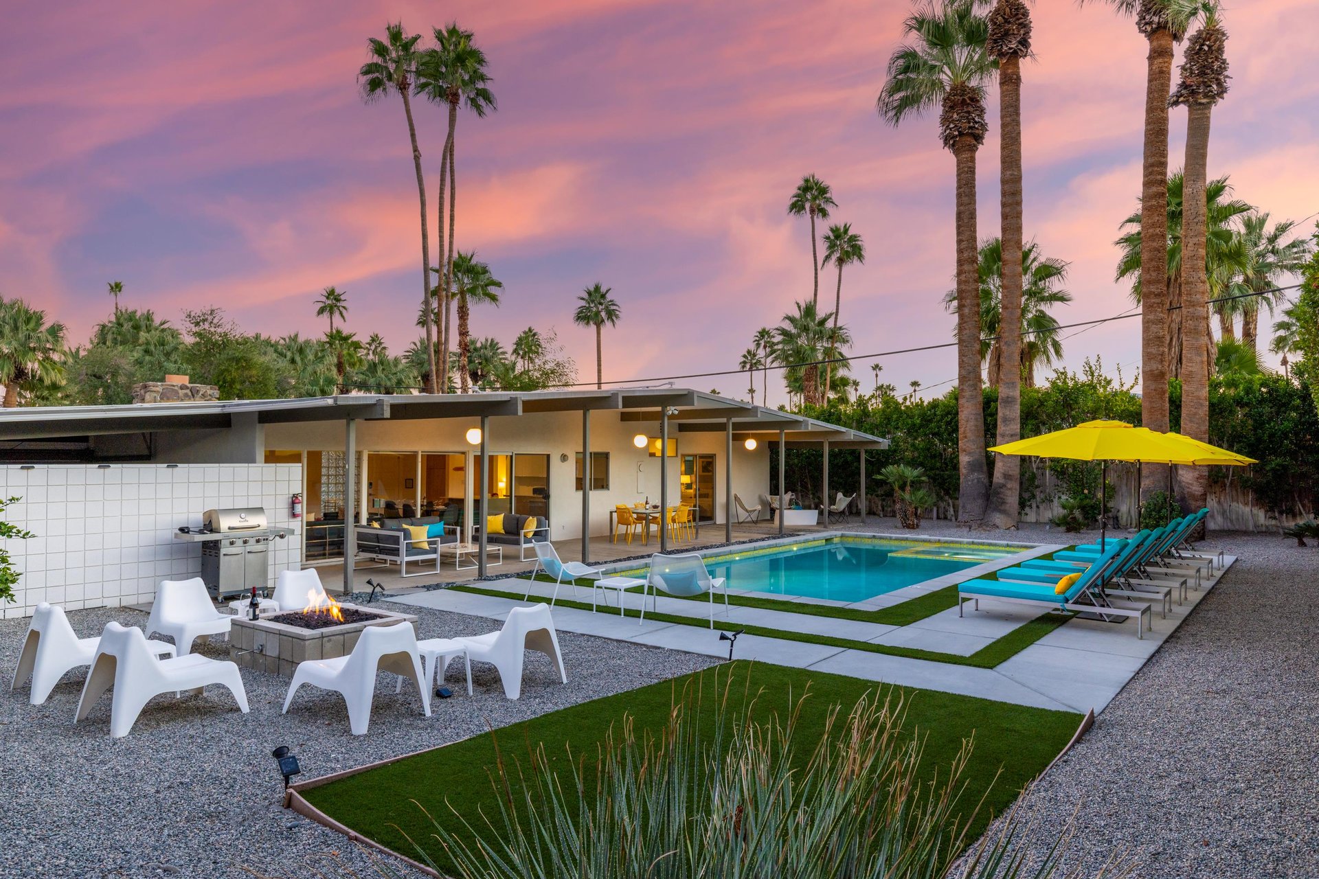 Modern desert retreat showcasing a stunning swimming pool area surrounded by towering palm trees against a vibrant sunset sky.