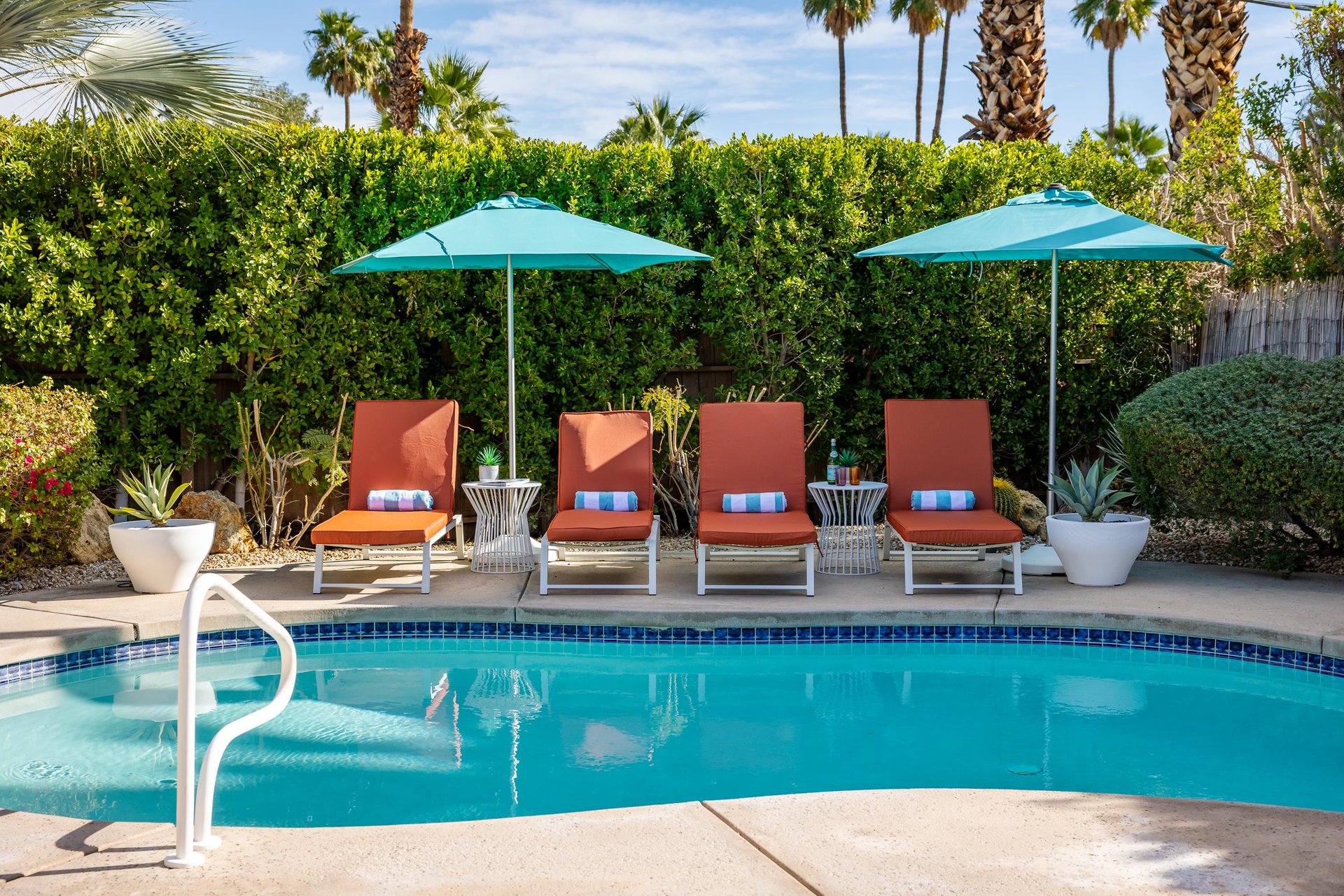 Lounge by your private pool on cushioned chairs under vibrant umbrellas, surrounded by lush palms and desert plants in this sunny oasis.
