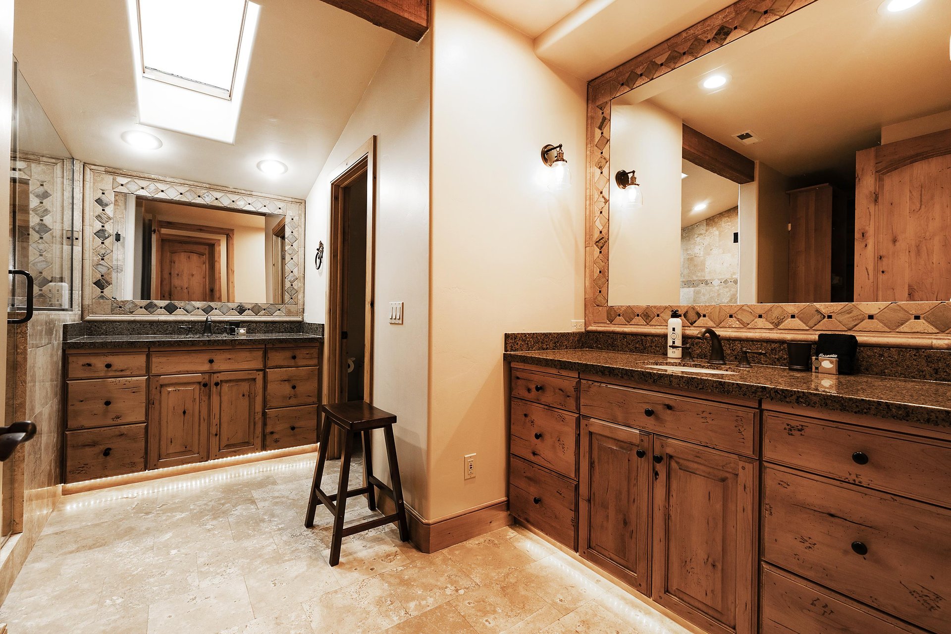 Ensuite bathroom with separate vanities with large mirrors, medium toned wooden cabinetry, and cream colored tiled floors.
