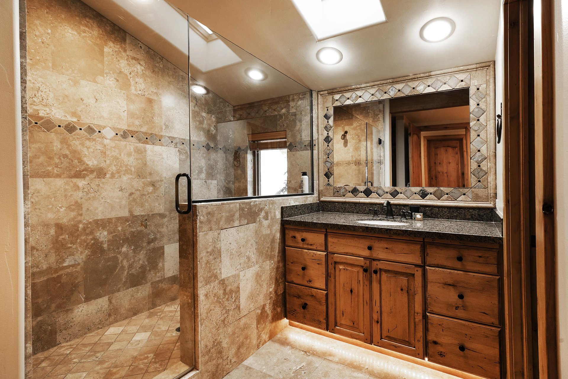 Cream colored tiled walkin shower with glass door, sky lights, medium wood vanity with large mirror and single sink.