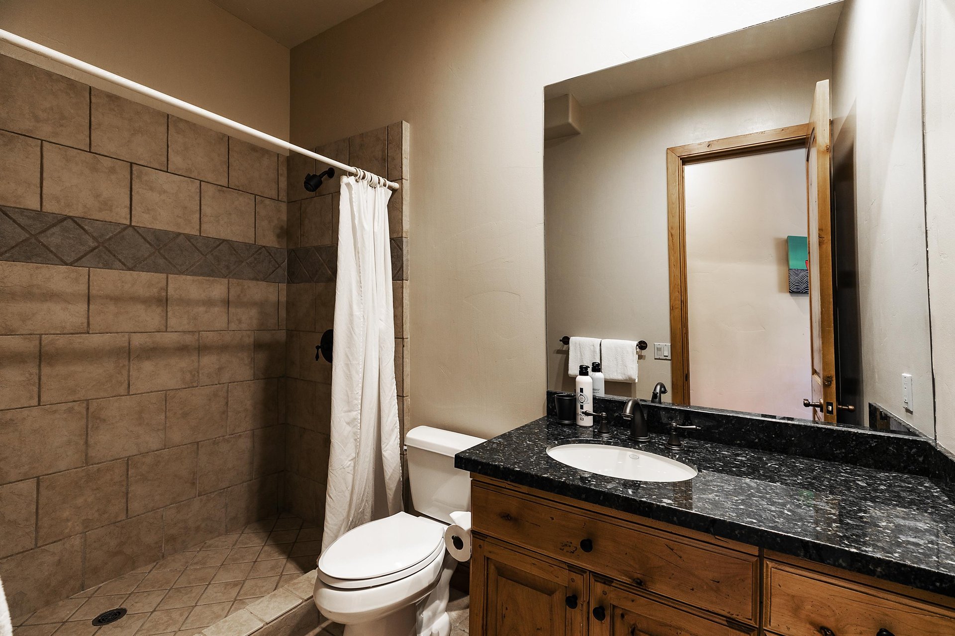Shower with cream colored tile and shower curtain, toilet, wooden vanity with black countertop, sink, and large mirror.