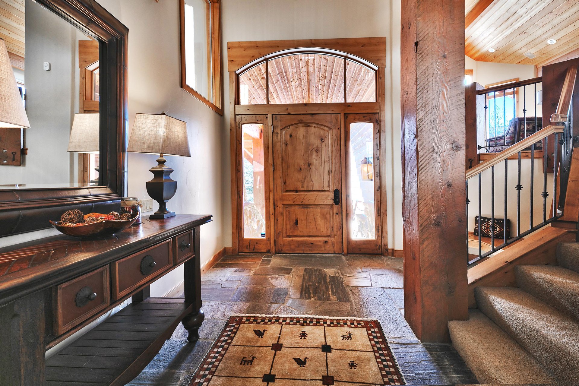 The Thistle Home entryway has a solid wood door with transom windows, slatestone floor, dark wood entry table and lamp.