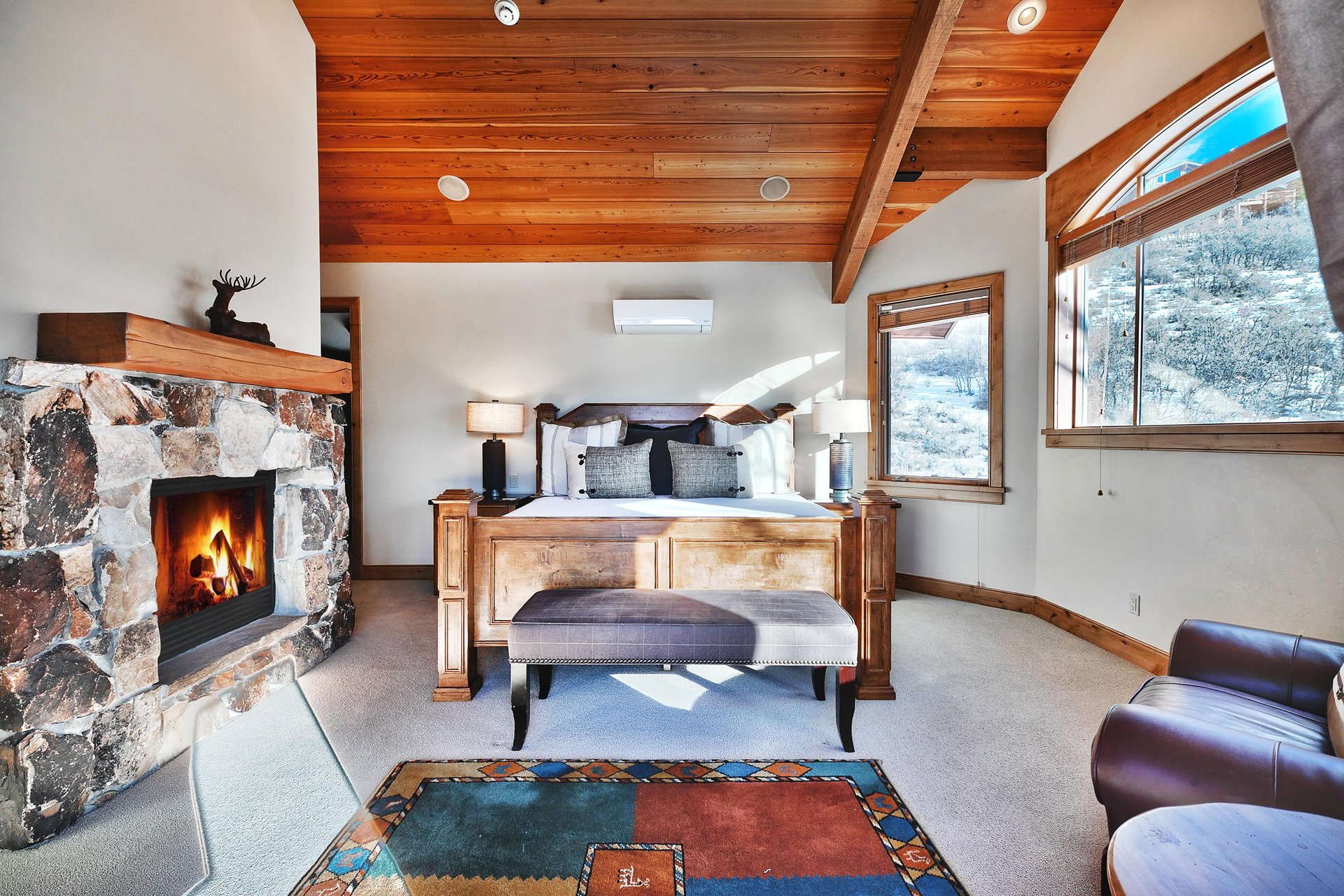 Four poster wooden king bed with grey upholstered bench at the end, a flagstone fireplace hearth and western inspired rug.