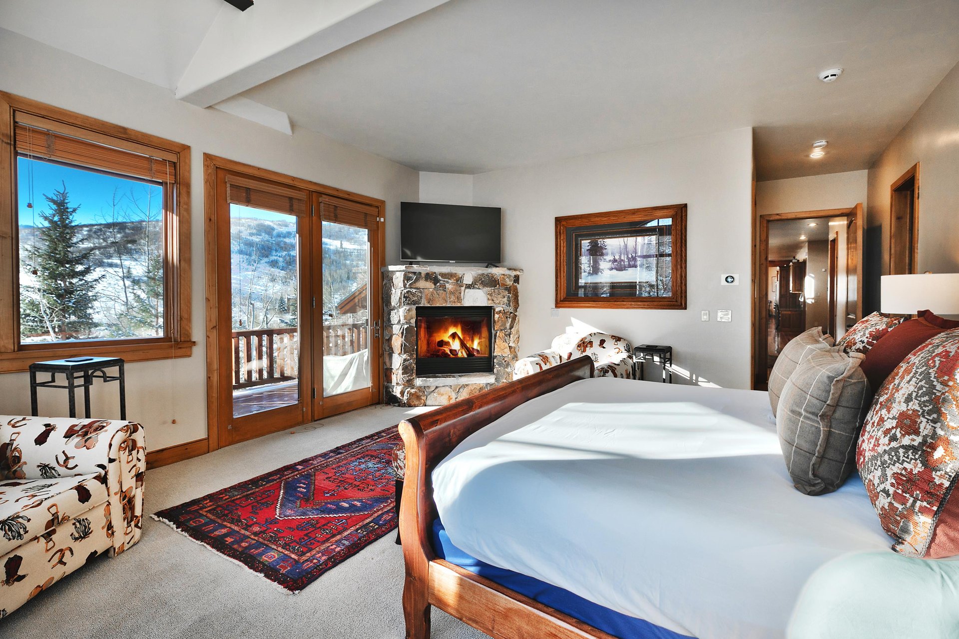 Wooden king sleigh bed with white linens, private patio glass doors, built in fireplace with stone surround and TV above.