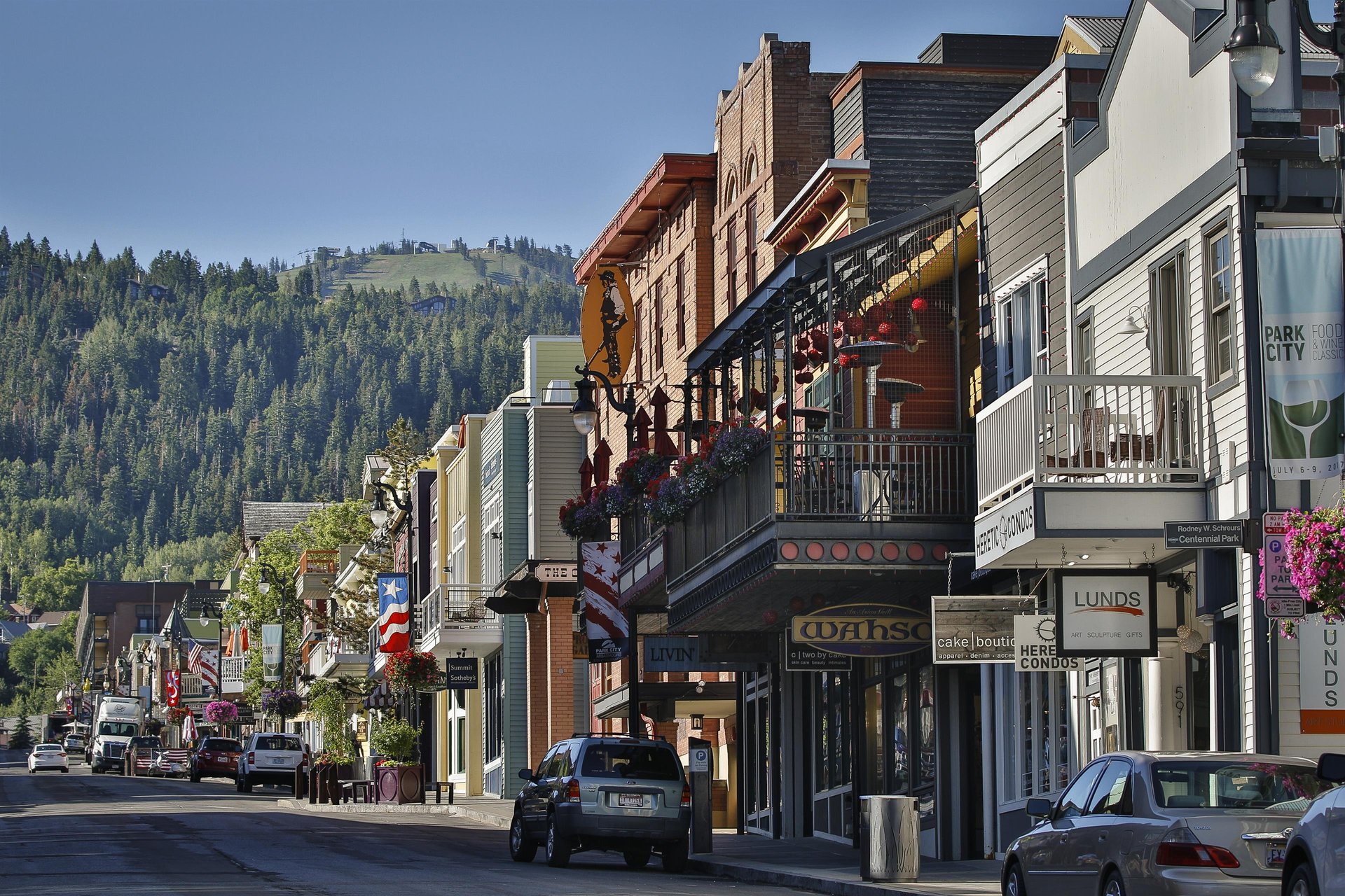 A charming mountain town main street lined with historic buildings, local shops, and eateries, set against a backdrop of lush forested hillsides and clear blue skies.