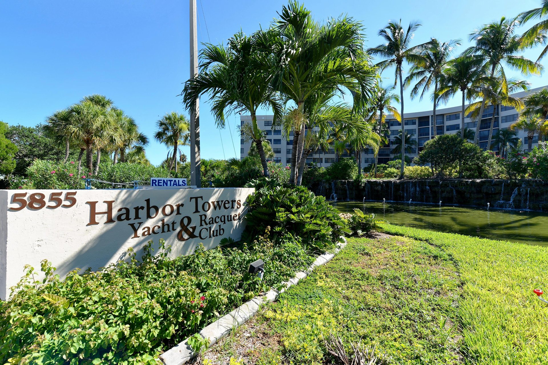Harbor Towers Yacht  Racquet Club entrance surrounded by lush tropical landscaping and towering palm trees under brilliant blue skies.