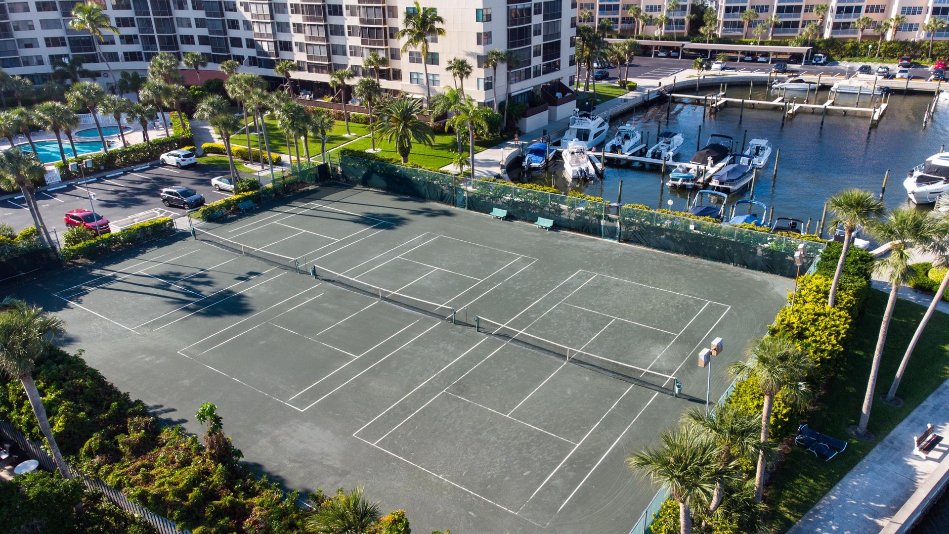 Aerial view of tennis courts surrounded by tropical landscaping with marina and residential buildings nearby.