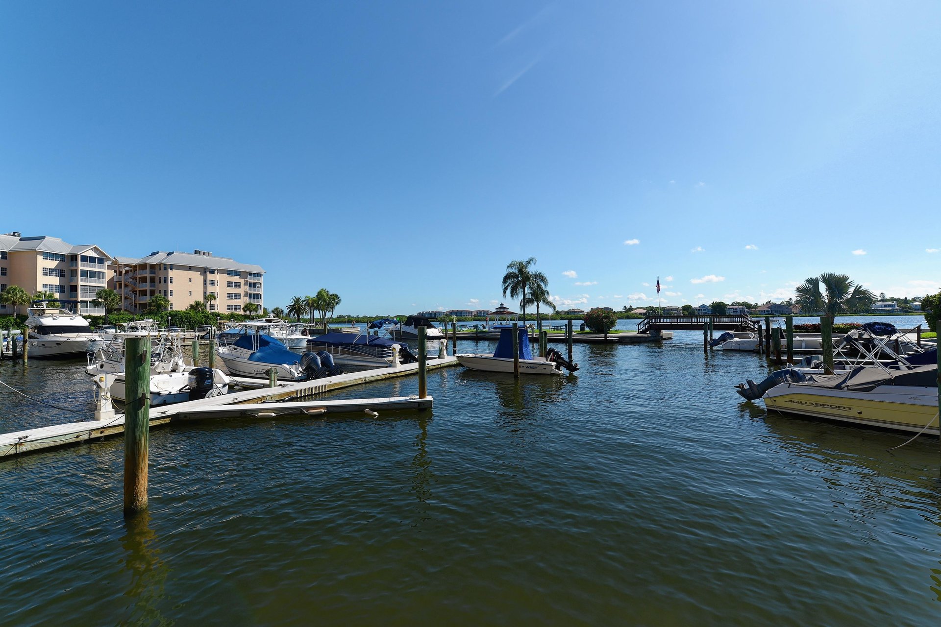 Marina with boats docked along waterfront, featuring palm trees and nearby buildings under clear blue skies.