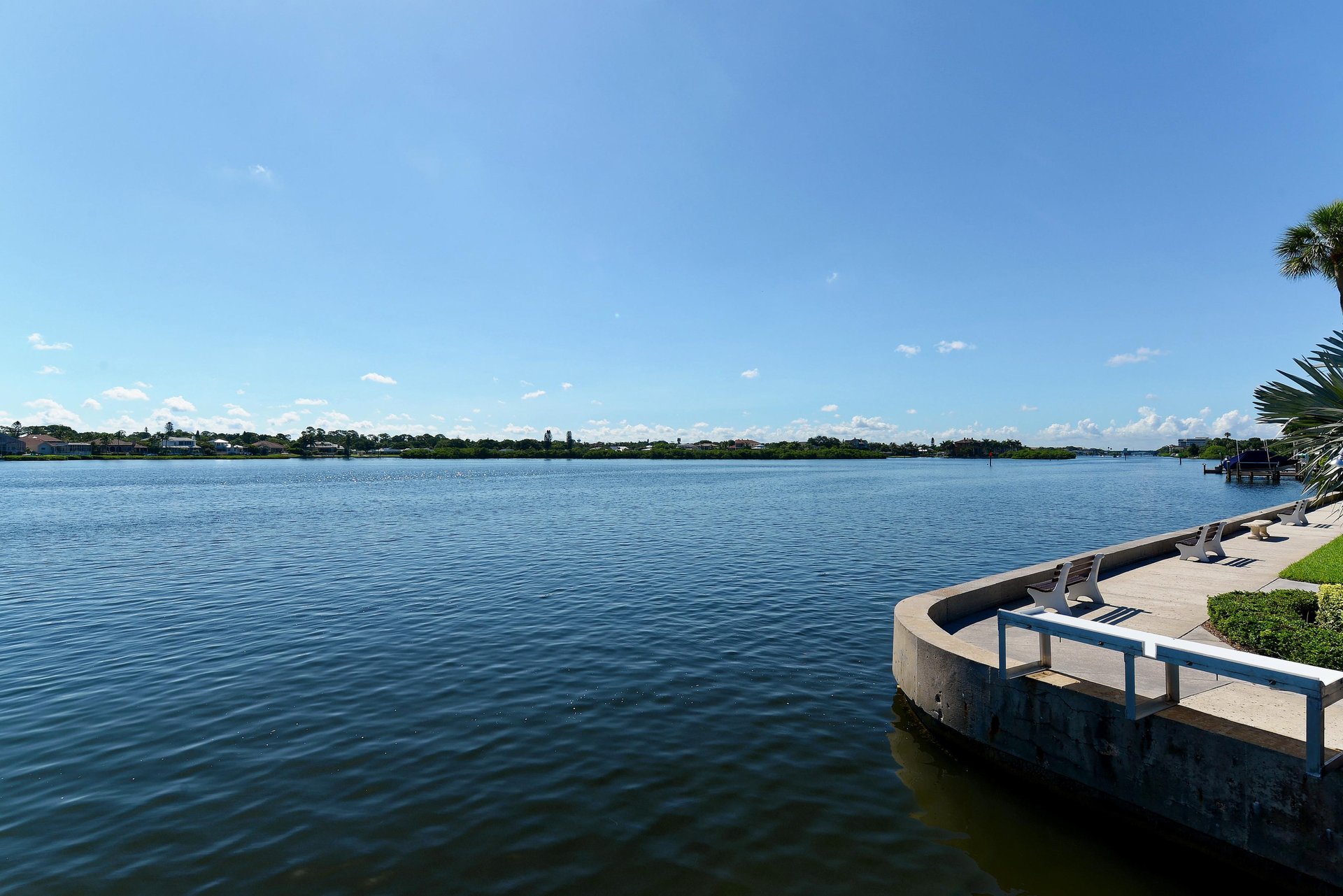 Waterfront seating area overlooking a peaceful lake with distant residential shoreline and blue sky.
