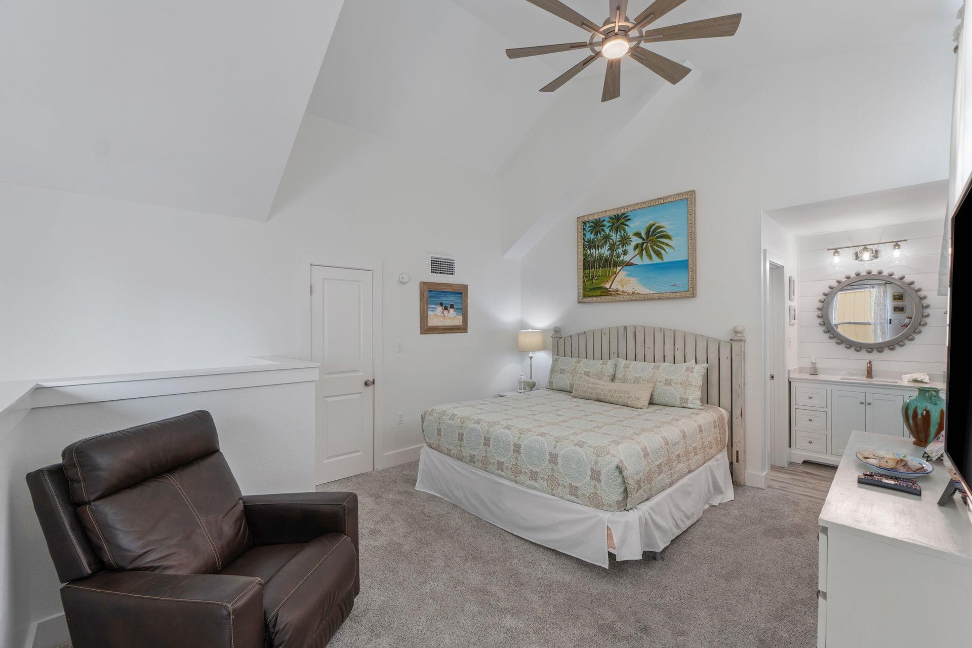 Relax in your serene bedroom sanctuary with coastal artwork, vaulted ceilings, and a cozy leather chair for unwinding after beach days.