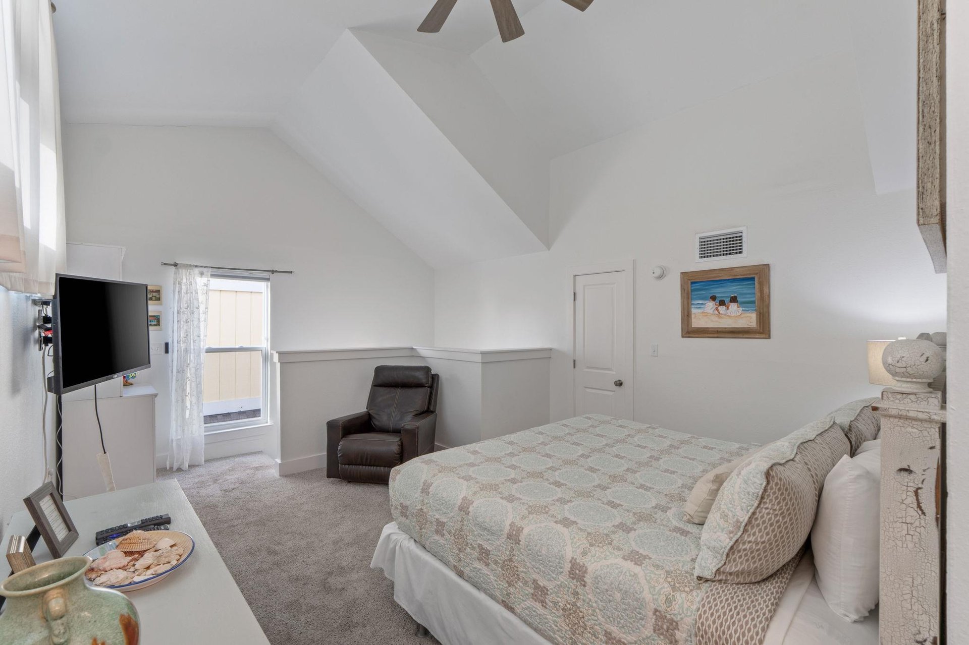 Relax in your spacious bedroom featuring vaulted ceilings, cozy seating, and coastal artwork that creates a peaceful retreat for restful nights.