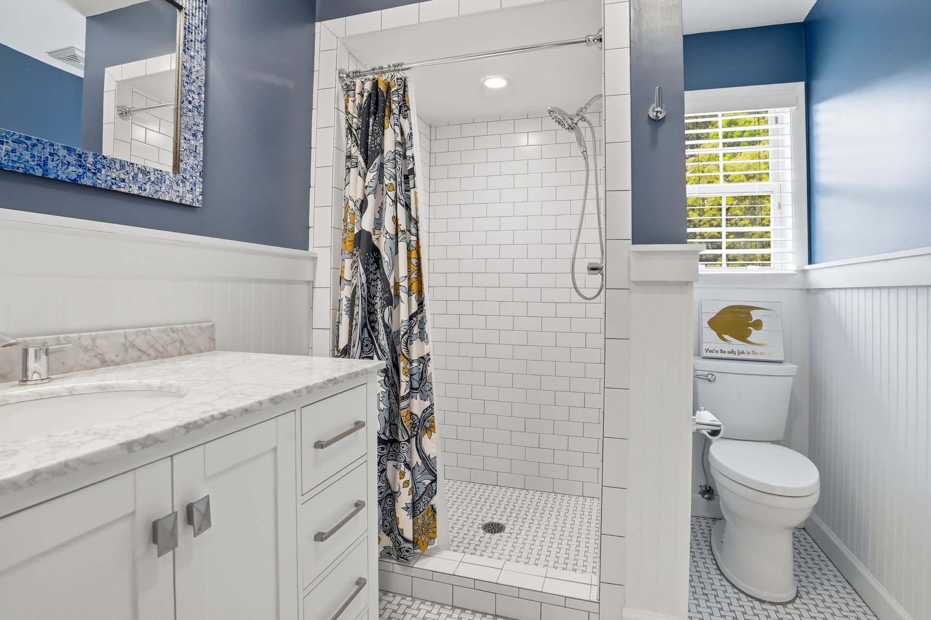 Navy accent wall, mosaicframed mirror, white vanity, and a subway tile tubshower — the hall bath matches the nautical rooms energy.
