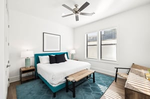 Rest peacefully in your bright bedroom featuring a plush teal bed, modern ceiling fan, and warm natural light streaming through oversized windows.