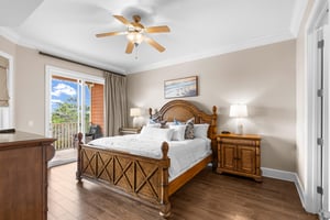 First floor king bedroom with tropical wood furnishings, ceiling fan, and private balcony overlooking lush landscaping.