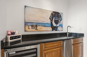 This stylish wet bar awaits with wine fridge, ice maker, sink, and charming dog artwork—perfect for evening cocktails and coastal conversations.