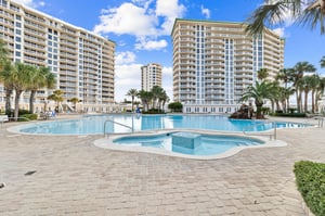 Modern resort towers frame a sparkling pool complex with integrated hot tub, surrounded by tropical palms and elegant stonework.
