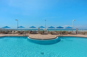 Sparkling pool with beachfront access featuring turquoise umbrellas and lounge chairs overlooking pristine sandy shores.