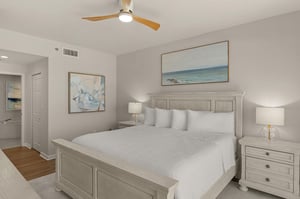 Bedroom 1 with king bed awaits with crisp white linens, coastal artwork, and gentle ceiling fan creating the perfect retreat.
