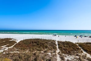 Pristine white sand beach stretches along turquoise waters under clear blue skies, with natural dune vegetation creating coastal pathways.