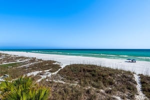 Pristine white sand beach stretches along emerald waters under clear blue skies, with natural dune vegetation and walking paths leading to the shore.
