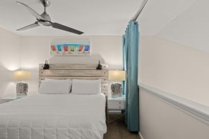 Unwind in your cozy coastal retreat with a plush bed, whimsical beach chair art, and soothing bedside lamps. The ceiling fan keeps you comfortable as ocean breezes drift through.