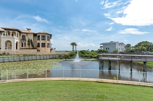 Vacation rental property surrounded by tranquil waterfront community with landscaped pond, fountain, walking bridge, and nearby neighboring homes under clear blue skies.