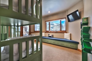 Bedroom with green twin bunk beds, wall mounted TV, window with snowy mountain views, window seat, and tree bookcase.