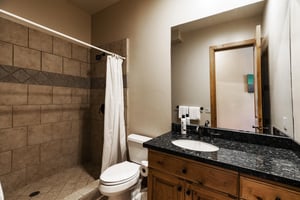 Shower with cream colored tile and shower curtain, toilet, wooden vanity with black countertop, sink, and large mirror.