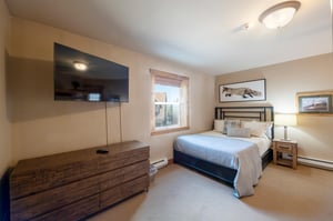 Rest easy in the comfortable guest bedroom at Resort Plaza 5002 — this Park City condo rental offers a clean, bright retreat with a flatscreen TV, rustic wood dresser, and charming mountain artwork f