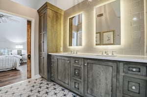 The primary en suite — double vanity, illuminated mirrors, gray cabinetry, and geometric tile floors.