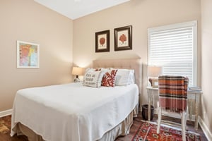 A warm and inviting guest room with a queen bed, plaid throw, framed art above the headboard, and a Persian rug underfoot.
