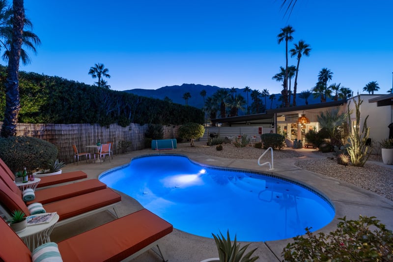 Stunning desert oasis featuring a glowing pool surrounded by palm trees and mountain views at twilight.