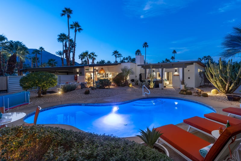 Modern desert oasis featuring illuminated swimming pool surrounded by palm trees and mountains at twilight.