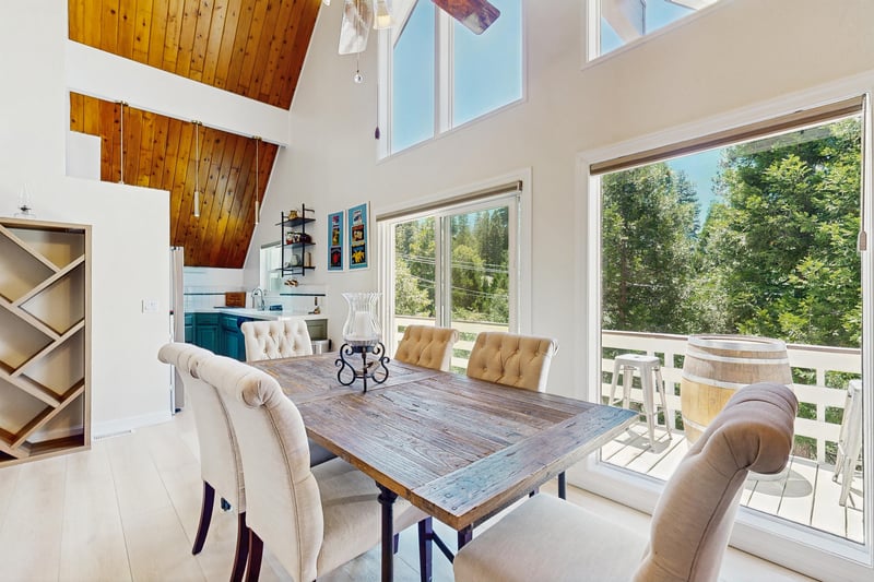 Dining area with vaulted ceilings, large windows, deck access, fireplace, and table and chairs for 6 guests.