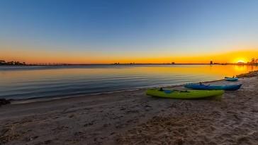 Paddleboards resting on sandy beach at sunrise beside smooth, glassy bay water with the sun rising on the horizon.