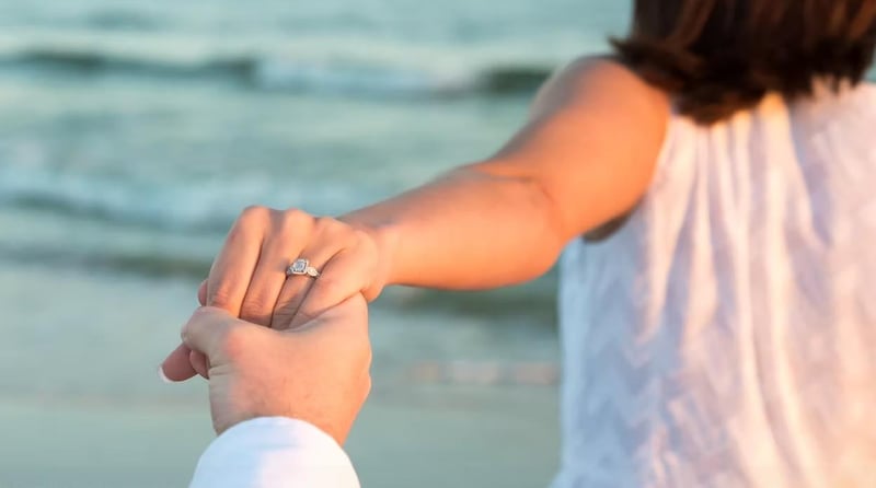 Closeup of a couple holding hands on the beach with a diamond engagement ring visible at sunset by the water.