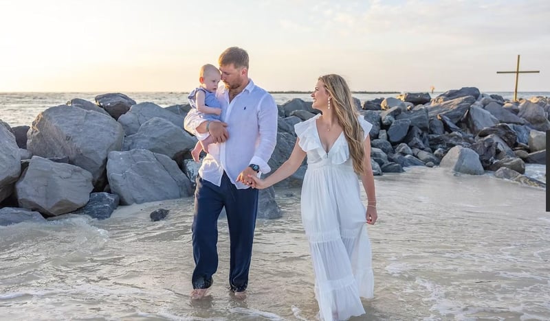 Parents walking barefoot through shallow water while holding their baby near coastal rocks at sunset during a beach portrait session.