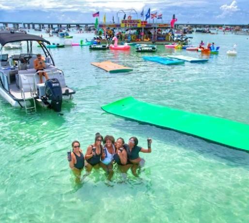Group of women standing in shallow water near a pontoon boat and floating mat at Crab Island with boats and a floating vendor in the background.