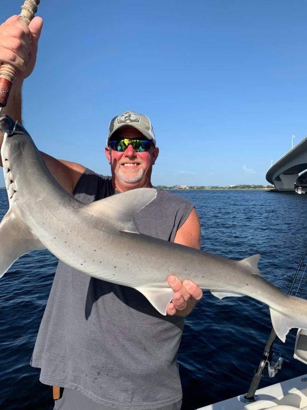 Man wearing sunglasses holding a shark on a boat near a coastal bridge during a guided shark fishing charter.