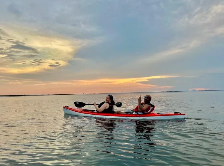 Two adults paddling a red tandem kayak on the waters of Pensacola Beach with clear blue skies.