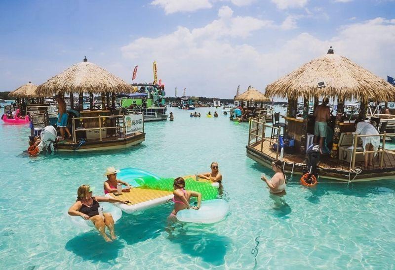 Multiple tiki boats anchored at Crab Island with people relaxing in shallow turquoise water.